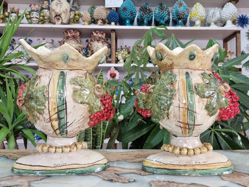 May include: Two ceramic vases with a crown-like top, decorated with green leaves, red berries, and a yellow and green striped pattern. The vases are on a white surface with a green and yellow border. The text "Tradizioni" is visible on the base of one vase.