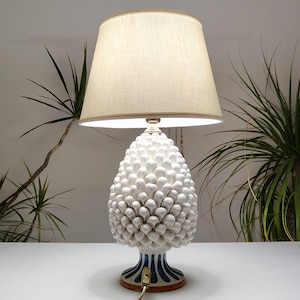 May include: A decorative table lamp with a white ceramic base shaped like a pinecone. The lamp has a beige fabric lampshade with a gold trim. The base features blue and white stripes. The lamp is on a white surface.