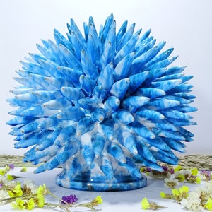 Pode incluir: Uma escultura de cerâmica azul e branca com uma forma redonda e espinhosa. A escultura é feita de muitas peças pequenas e pontiagudas que são dispostas em um padrão circular. A escultura está sentada em uma superfície branca.