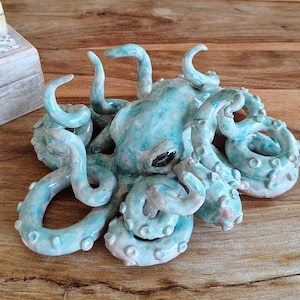 May include: A ceramic octopus sculpture in shades of turquoise and pink. The tentacles curl and feature small, textured suckers. The sculpture has a glossy finish and sits on a wooden surface.