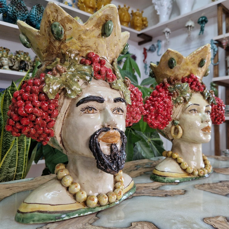 May include: Two ceramic sculptures of people's heads with crowns, red berries, and green leaves. The sculptures are painted in shades of white, yellow, green, and red.