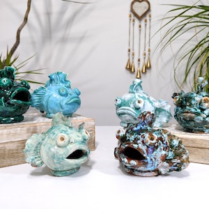 Può includere: Una collezione di sei sculture di pesci in ceramica in vari colori, tra cui verde acqua, blu e marrone. Ogni pesce ha la bocca aperta e dettagli, posti su blocchi di legno. Sculture decorative e artistiche.