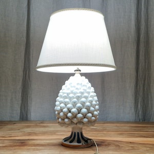 May include: White ceramic table lamp with a pine cone shaped base and a white fabric lampshade. The base has blue and white stripes.