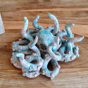 May include: A ceramic octopus sculpture in shades of blue, teal, and pink. The octopus has multiple curled tentacles and a detailed head with dark eyes. The sculpture is placed on a wooden surface.