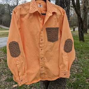 Può includere: Camicia arancione con bottoni con colletto, tasca e toppe sui gomiti ricamate. La camicia presenta un piccolo disegno ricamato sul colletto e sui polsini e una tasca strutturata. Le toppe sui gomiti sono di colore marrone scuro.
