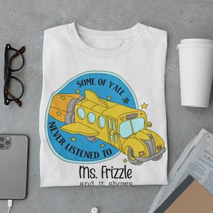 May include: A white t-shirt featuring a yellow school bus flying through space with the text "Some of y'all never listened to Ms. Frizzle and it shows."