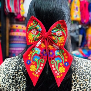 May include: A red bow with colorful floral embroidery. The bow is tied in the back of a person's hair.
