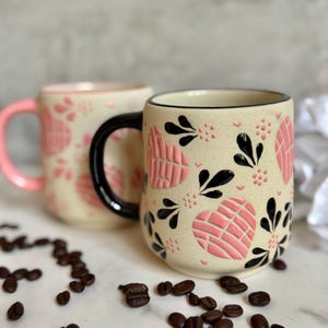 May include: Two ceramic mugs with a beige base, adorned with pink heart-shaped designs and black floral accents. One mug has a pink handle, the other a black handle. Coffee beans are scattered around the mugs.