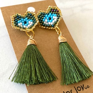 May include: A pair of green tassel earrings with a beaded heart-shaped charm featuring a blue evil eye design. The earrings are on a brown card with the text "color joy co."