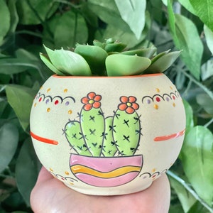 May include: A small, round, cream-colored ceramic planter with a painted cactus design. The cactus is green with orange flowers and is potted in a pink pot with yellow stripes. The planter has a white background with orange, red, and purple dots and lines.