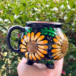 May include: Black ceramic mug with a handle and three legs. The mug is decorated with yellow and orange sunflowers and green leaves on a black background.