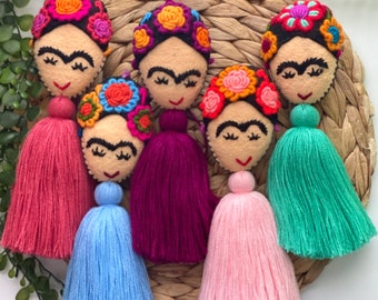 May include: Six colorful tassel keychains with embroidered felt faces. Each keychain features a different colored tassel and a felt face with a black unibrow and eyelashes. The faces are decorated with colorful flower embroidery.