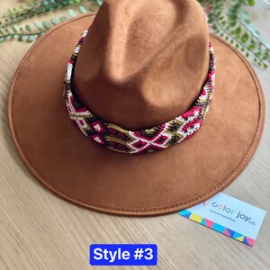 May include: A brown suede fedora hat with a colourful woven band. The band features geometric patterns in red, white, gold, and black. The hat is labelled "Style #3" and "color joy co."