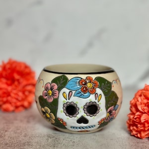 May include: A cream-coloured ceramic bowl with a Day of the Dead sugar skull design. The skull has black eye sockets, a pink nose, and teeth. Colourful flowers and green leaves surround the skull. Two orange flowers are on either side of the bowl.