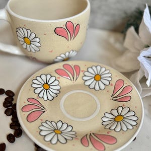 May include: A beige ceramic teacup and saucer set decorated with white daisies and pink accents. The cup has a handle and the saucer has a central indentation. Coffee beans are scattered around the set.