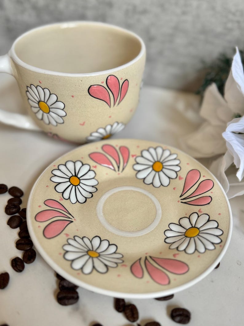 May include: A beige ceramic teacup and saucer set decorated with white daisies and pink accents. The cup has a handle and the saucer has a central indentation. Coffee beans are scattered around the set.