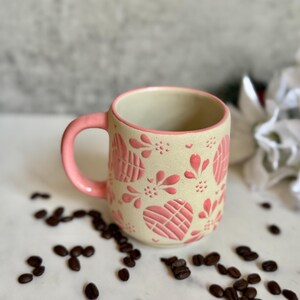 May include: A ceramic mug with a pink handle and rim, decorated with pink heart and floral patterns. The mug sits on a white surface scattered with coffee beans. The mug's exterior is a cream color.