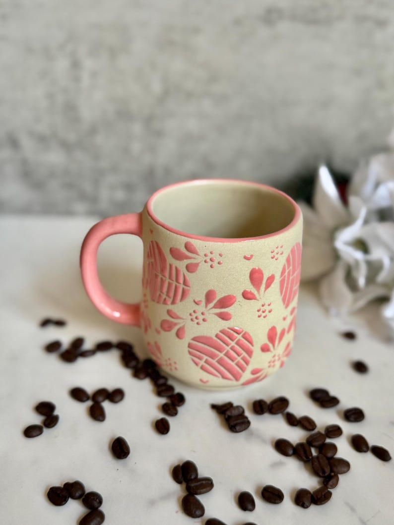 May include: A ceramic mug with a pink handle and rim, decorated with pink heart and floral patterns. The mug sits on a white surface scattered with coffee beans. The mug's exterior is a cream color.