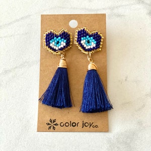 May include: A pair of blue tassel earrings with gold-toned evil eye charms. The charms are made of blue, white, and gold beads. The earrings are on a brown card with the text "color joy co."