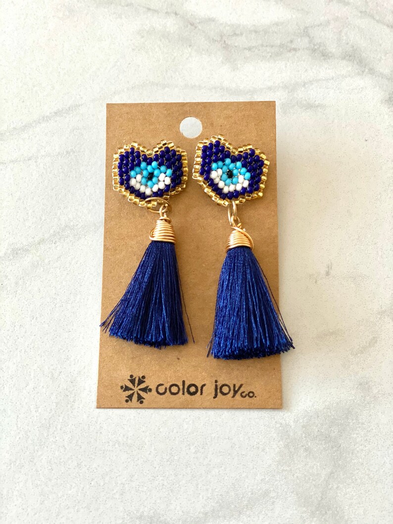 May include: A pair of blue tassel earrings with gold-toned evil eye charms. The charms are made of blue, white, and gold beads. The earrings are on a brown card with the text "color joy co."