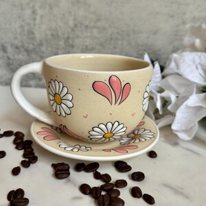 May include: A beige ceramic teacup and saucer set decorated with white daisies and pink accents. The cup has a curved handle and the saucer is round. Coffee beans are scattered around the set.