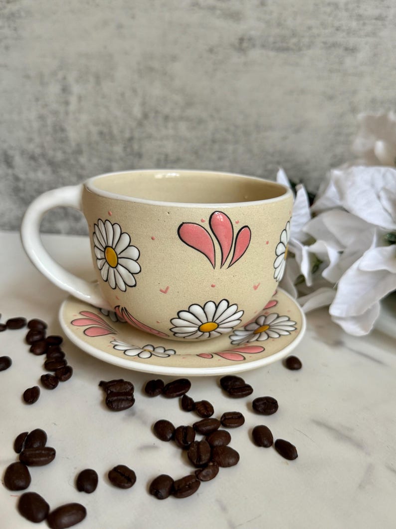 May include: A beige ceramic teacup and saucer set decorated with white daisies and pink accents. The cup has a curved handle and the saucer is round. Coffee beans are scattered around the set.