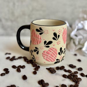 May include: A ceramic coffee mug with a beige background, black handle, and rim. The mug is decorated with pink heart shapes and black floral designs. Coffee beans are scattered around the mug.