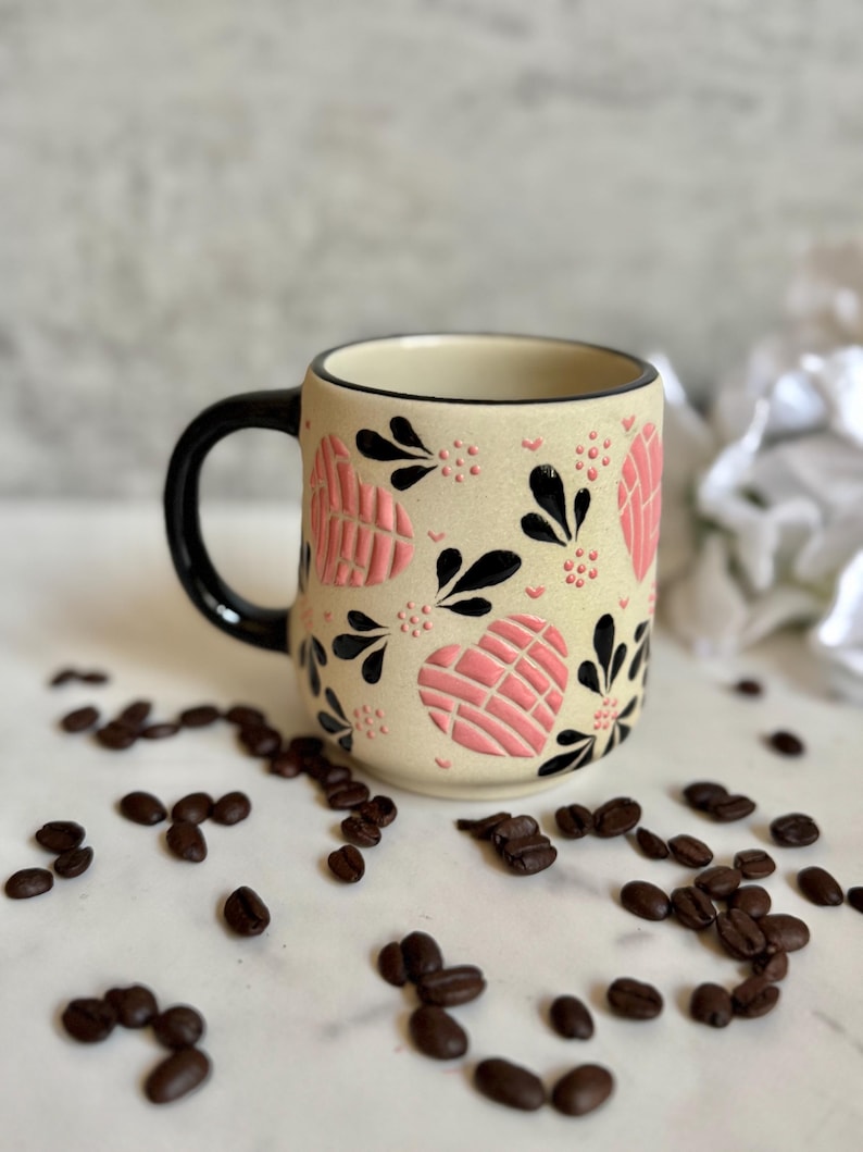 May include: A ceramic coffee mug with a beige background, black handle, and rim. The mug is decorated with pink heart shapes and black floral designs. Coffee beans are scattered around the mug.