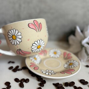 May include: A beige ceramic teacup and saucer set decorated with white daisies and pink petal accents. The cup has a white handle. Coffee beans are scattered around the set, suggesting a coffee or tea theme.