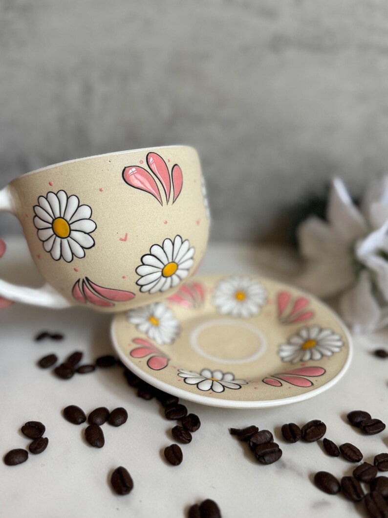 May include: A beige ceramic teacup and saucer set decorated with white daisies and pink petal accents. The cup has a white handle. Coffee beans are scattered around the set, suggesting a coffee or tea theme.