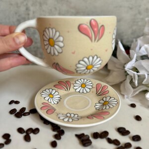May include: A beige ceramic coffee cup and saucer set decorated with white daisies and pink accents. The cup has a large handle and the saucer is round. Coffee beans are scattered around the set.