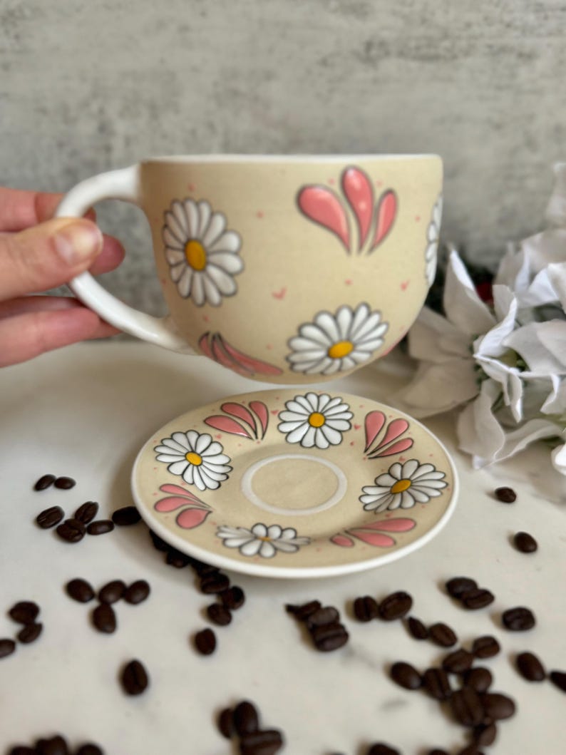 May include: A beige ceramic coffee cup and saucer set decorated with white daisies and pink accents. The cup has a large handle and the saucer is round. Coffee beans are scattered around the set.