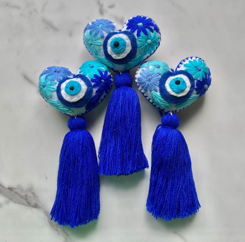 May include: Three blue and white heart-shaped ornaments with embroidered flowers and a blue evil eye charm. Each ornament has a blue tassel.