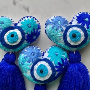 May include: Three blue and white felt heart-shaped ornaments with embroidered flowers and a blue evil eye charm. Each ornament has a blue tassel.