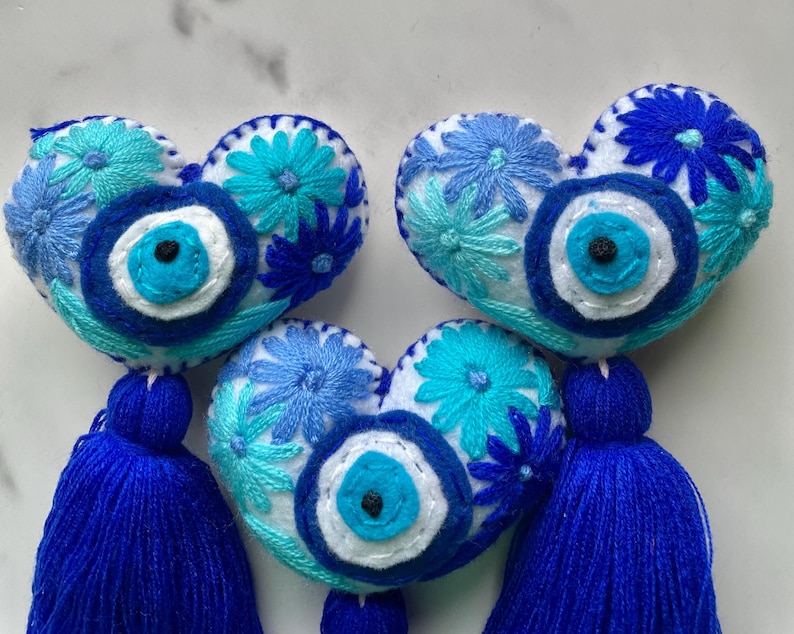 May include: Three blue and white felt heart-shaped ornaments with embroidered flowers and a blue evil eye charm. Each ornament has a blue tassel.