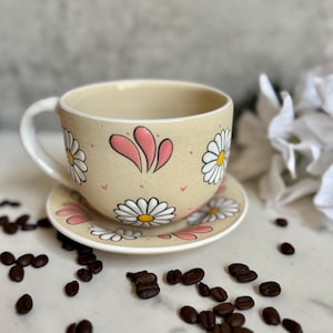 May include: A beige ceramic teacup and saucer set decorated with white daisies and pink accents. The cup has a white handle and sits on a matching saucer. Coffee beans are scattered around the set, suggesting a coffee or tea theme.