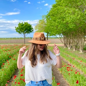 May include: A woman wearing a white shirt and blue jeans stands in a field of red poppies. She is wearing a brown hat with a patterned band.
