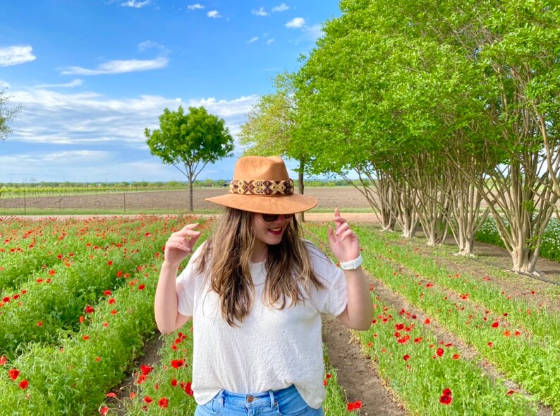 May include: A woman wearing a white shirt and blue jeans stands in a field of red poppies. She is wearing a brown hat with a patterned band.