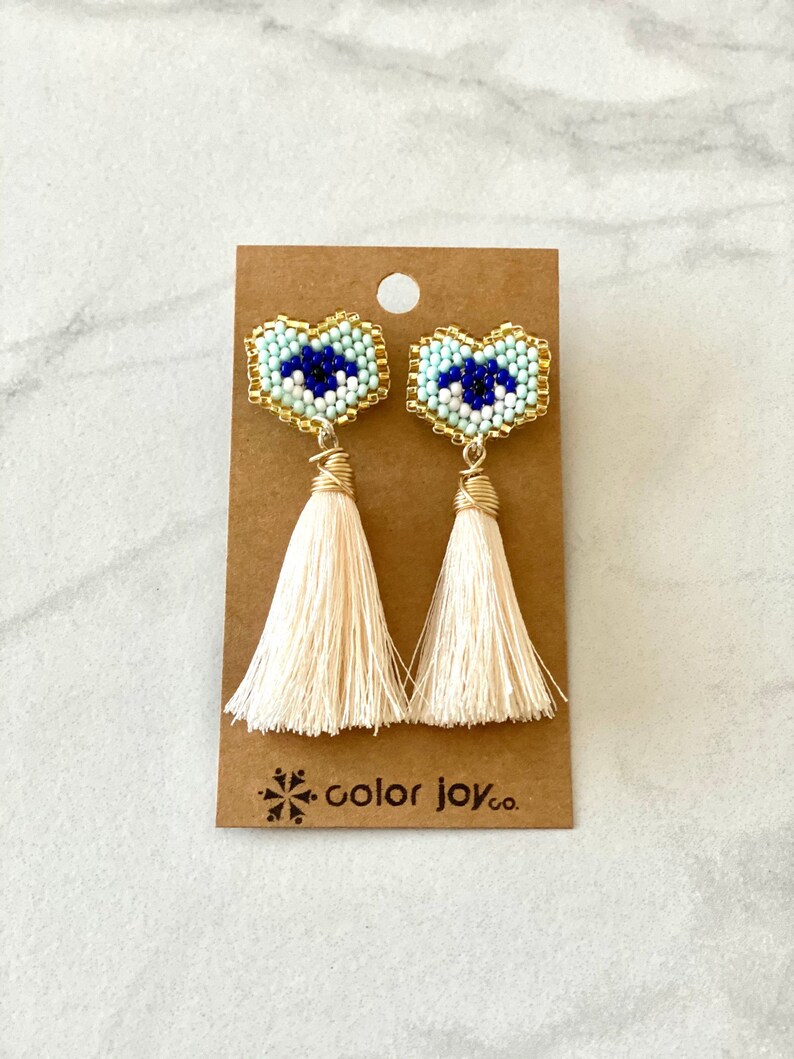 May include: A pair of white tassel earrings with blue and white beaded evil eye charms. The earrings are on a brown card with the text "color joy co."