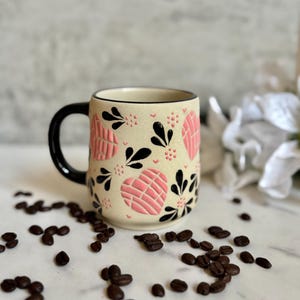 May include: Cream-colored ceramic mug with a black handle and rim, adorned with pink heart-shaped designs and black floral accents. Coffee beans are scattered around the base of the mug, creating a warm, inviting aesthetic.
