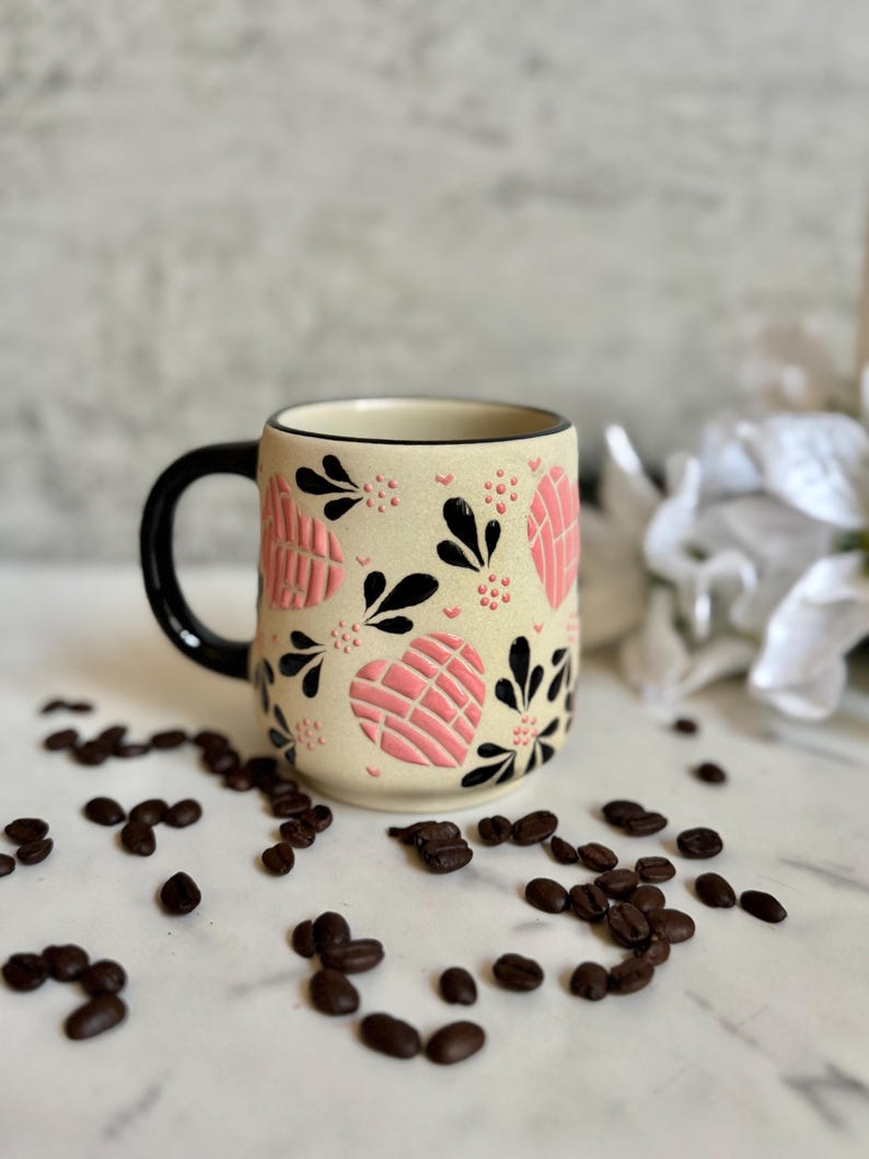 May include: Cream-colored ceramic mug with a black handle and rim, adorned with pink heart-shaped designs and black floral accents. Coffee beans are scattered around the base of the mug, creating a warm, inviting aesthetic.