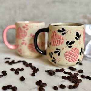 May include: Two ceramic mugs with heart and floral designs. One mug has a pink handle and the other has a black handle. The mugs are beige with pink heart patterns and black floral accents. Coffee beans are scattered around the mugs.