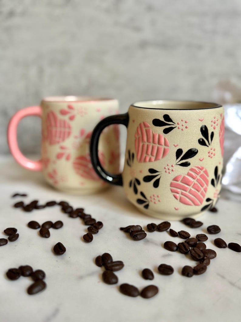 May include: Two ceramic mugs with heart and floral designs. One mug has a pink handle and the other has a black handle. The mugs are beige with pink heart patterns and black floral accents. Coffee beans are scattered around the mugs.