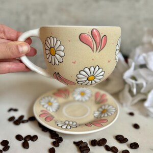 May include: A beige ceramic teacup and saucer set decorated with white daisies and pink accents. The cup has a white handle and the saucer is round. Coffee beans are scattered around the set.
