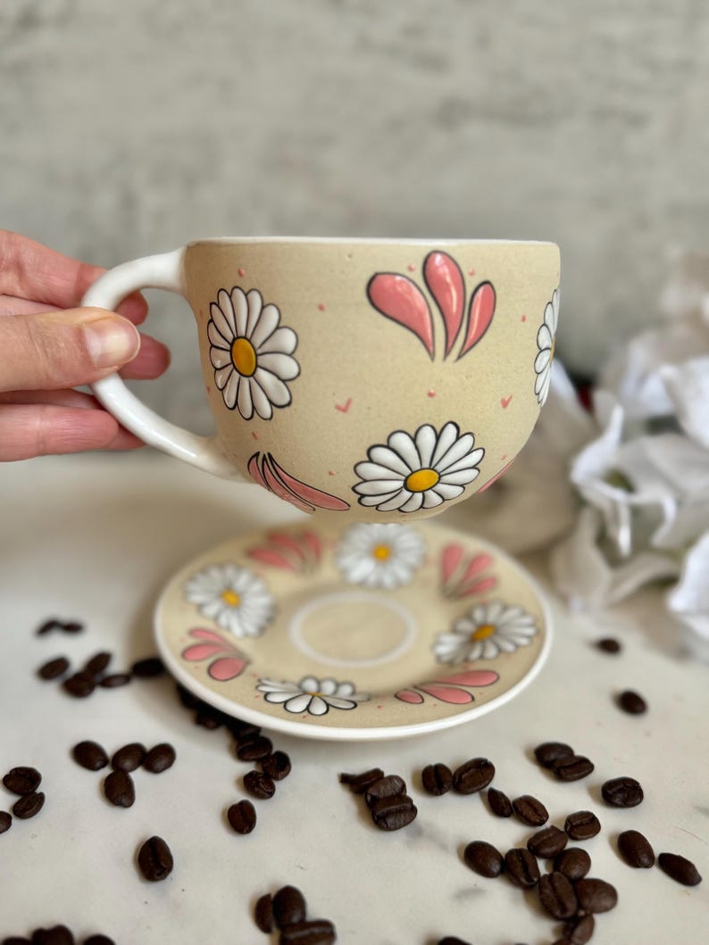 May include: A beige ceramic teacup and saucer set decorated with white daisies and pink accents. The cup has a white handle and the saucer is round. Coffee beans are scattered around the set.