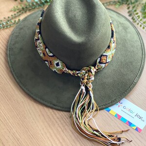 May include: A green felt fedora hat with a woven, multi-coloured band and tassel. The band is made of green, white, black, and gold threads. The tassel is made of the same colours. The hat is sitting on a wooden surface. The text "color joy co." is visible on a tag in the lower right corner of the image.