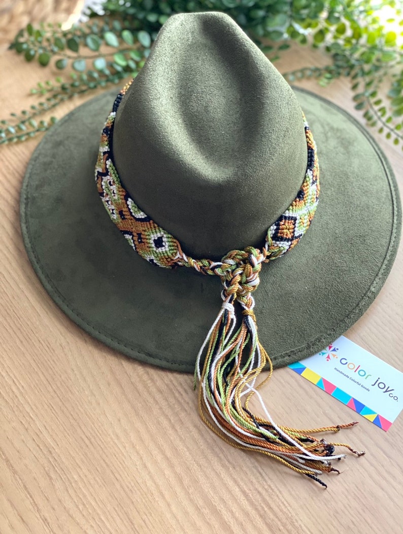 May include: A green felt fedora hat with a woven, multi-coloured band and tassel. The band is made of green, white, black, and gold threads. The tassel is made of the same colours. The hat is sitting on a wooden surface. The text "color joy co." is visible on a tag in the lower right corner of the image.