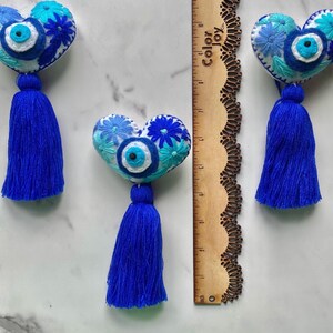 May include: Three white felt heart-shaped ornaments with blue and white embroidery, including a blue evil eye charm, and a blue tassel hanging from each.
