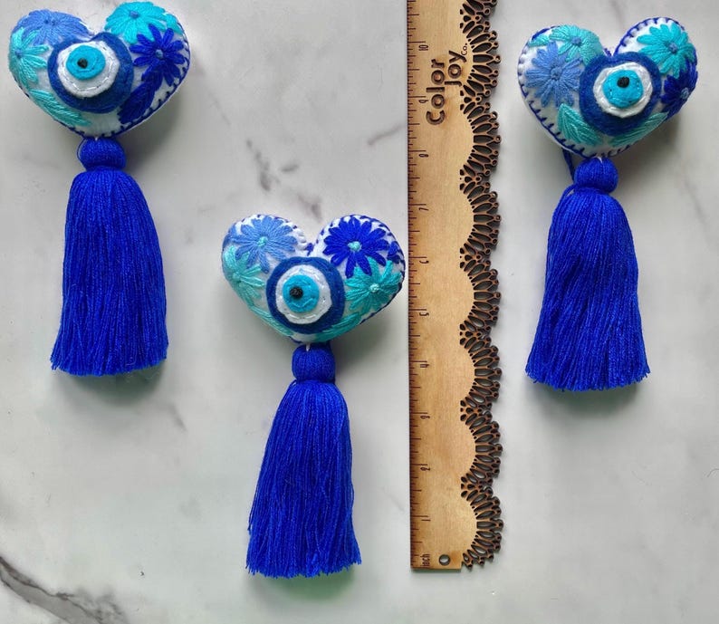 May include: Three white felt heart-shaped ornaments with blue and white embroidery, including a blue evil eye charm, and a blue tassel hanging from each.