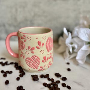 May include: A ceramic coffee mug with a cream-colored base and a pink handle. The mug is decorated with pink heart and floral patterns. Coffee beans are scattered around the mug on a white surface.